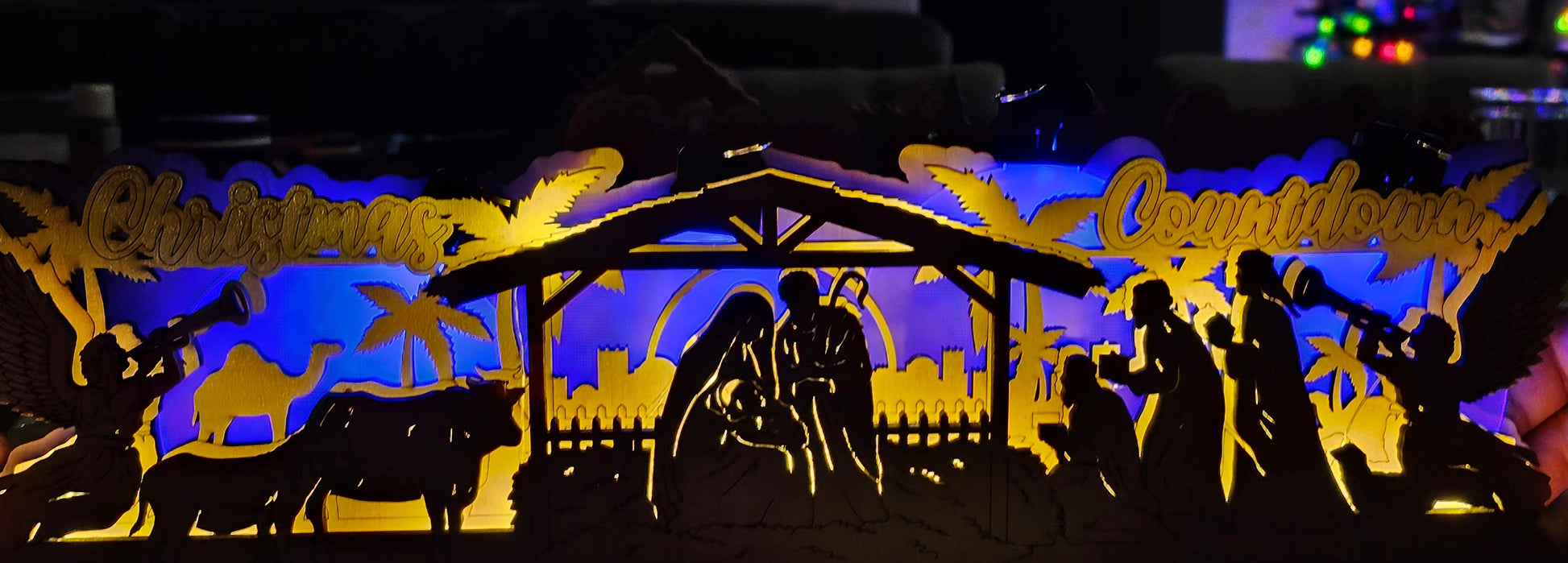 Nativity scene cutout with blue and yellow lighting in a room with a TV and Christmas tree.