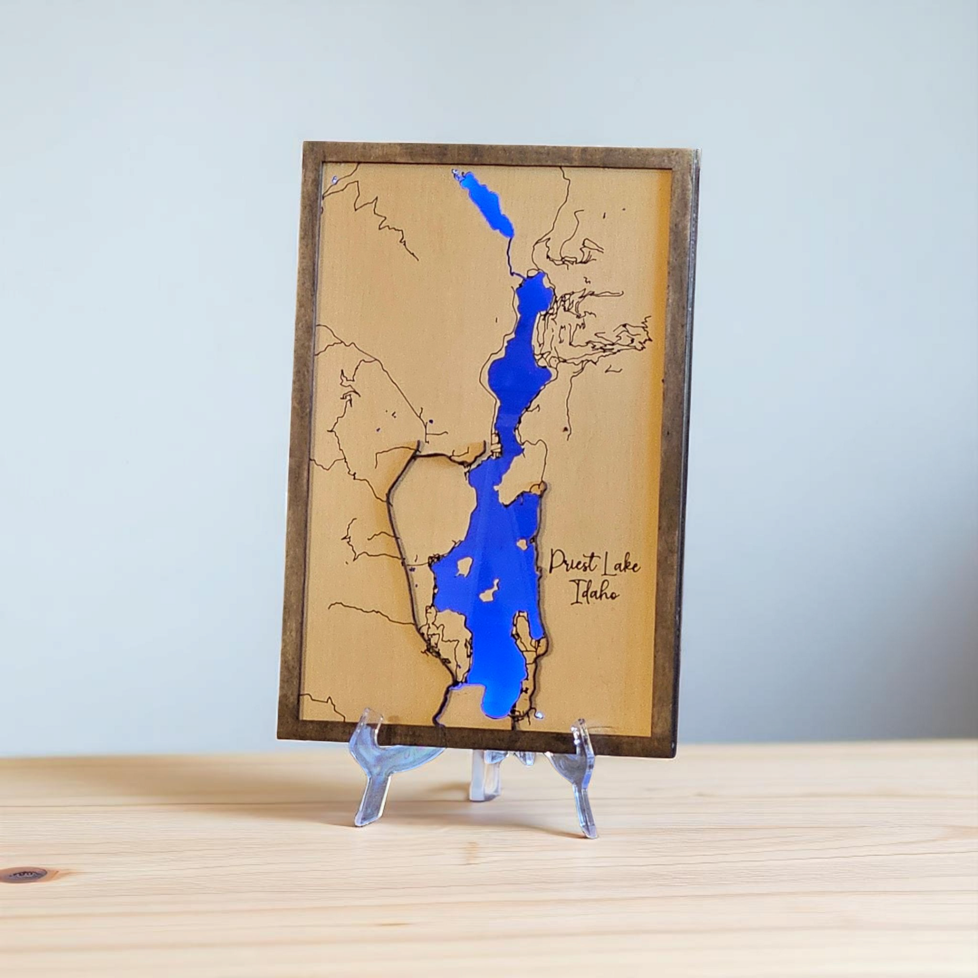 Wooden map of Priest Lake, Idaho with blue resin detailing on a stand against a light gray background.