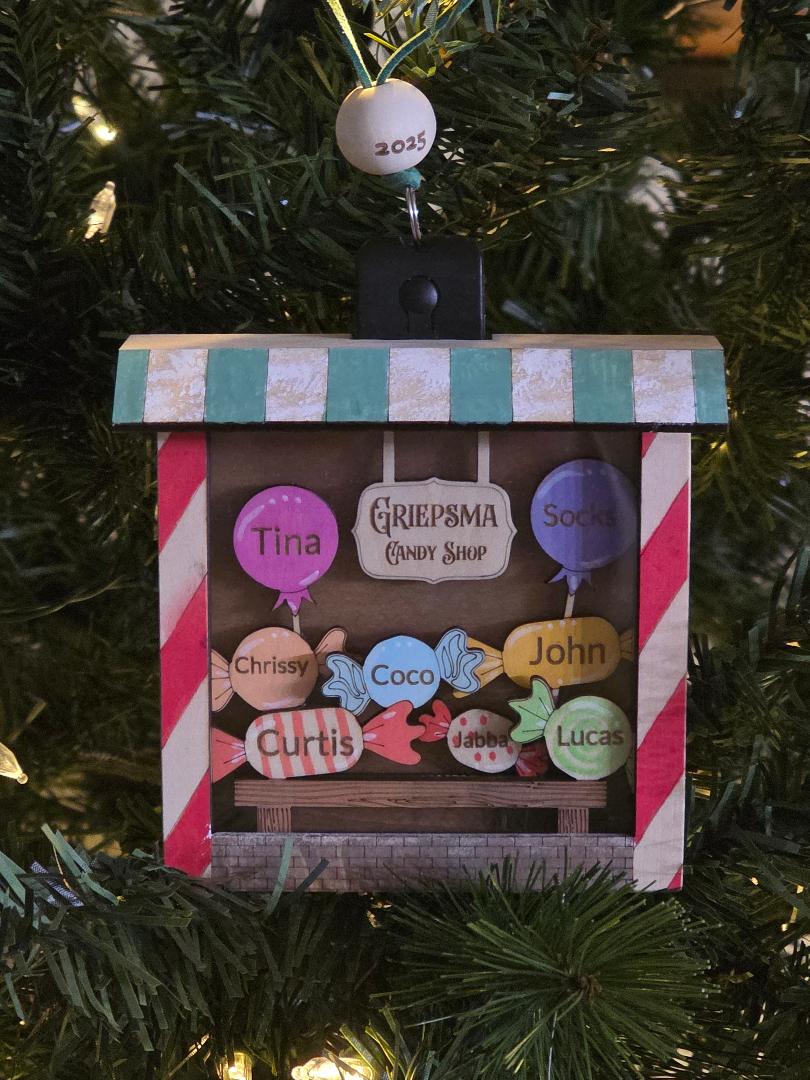 Decorative Christmas ornament with names on a candy shop sign against a Christmas tree background.